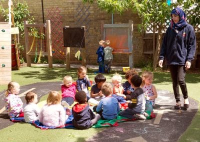 Children in the garden on Cambridge nursery, sitting in a group and listening to their teacher