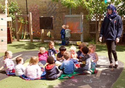 Children in the garden on Cambridge nursery, sitting in a group and listening to their teacher